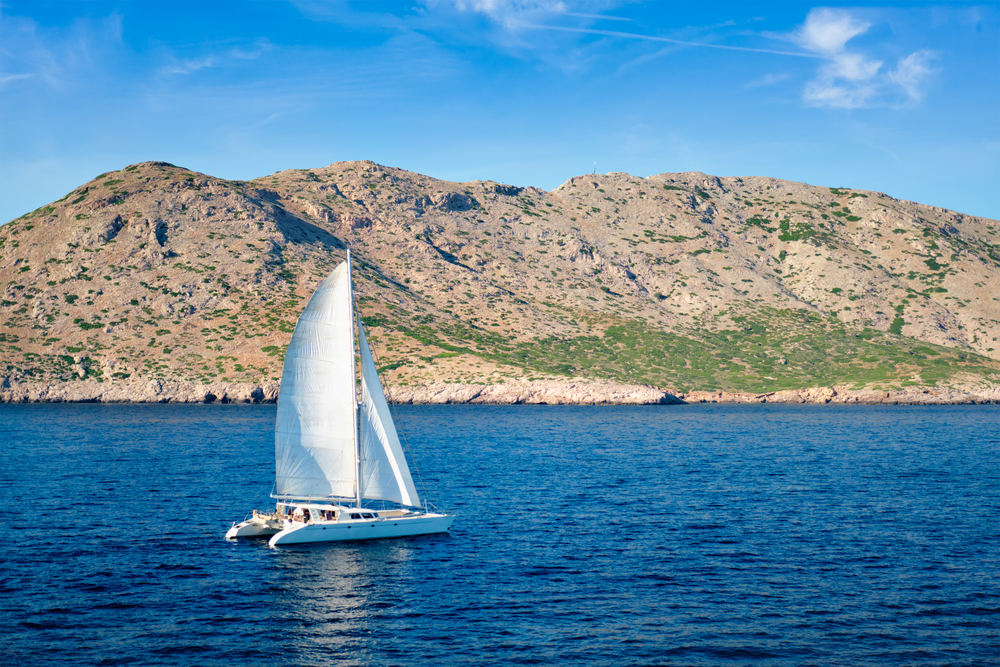 Catamaran sailing through blue waters in Greece with people sunbathing, BBQ on deck, and distant islands in view