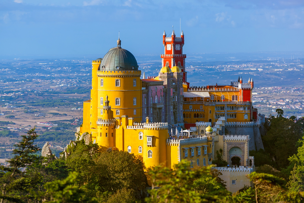 Colorful view of Pena Palace sitting atop a forested hill in Sintra, Portugal, with bright red and yellow towers against a blue sky.