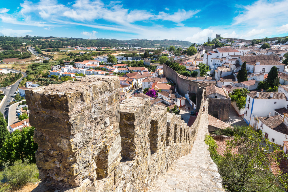 View of the medieval town of Óbidos, with whitewashed houses, a stone castle wall, and red-tiled rooftops under a sunny sky.