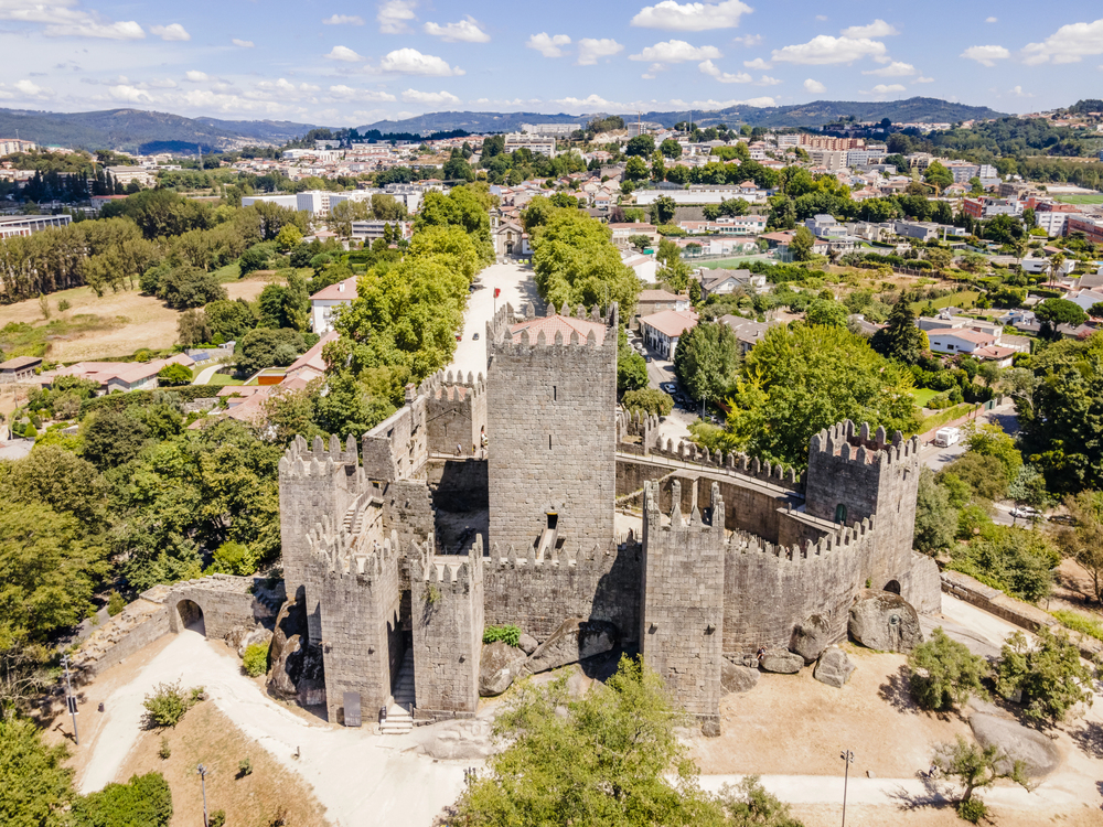 View of Guimarães Castle surrounded by green trees, with stone towers and flags waving under a clear blue sky.